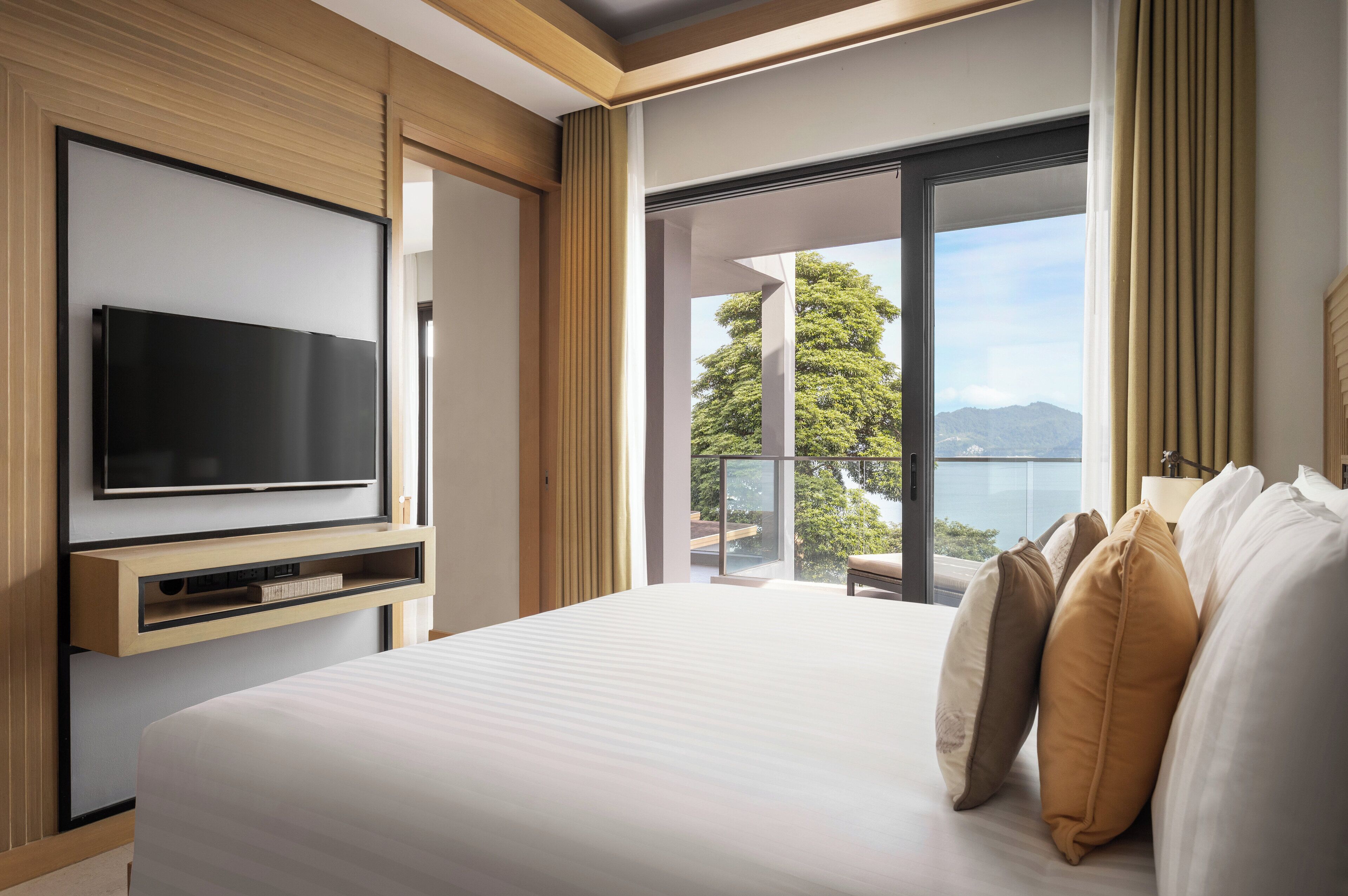 A hotel room with a large bed, a flat-screen TV, and a balcony with a view of trees and mountains.