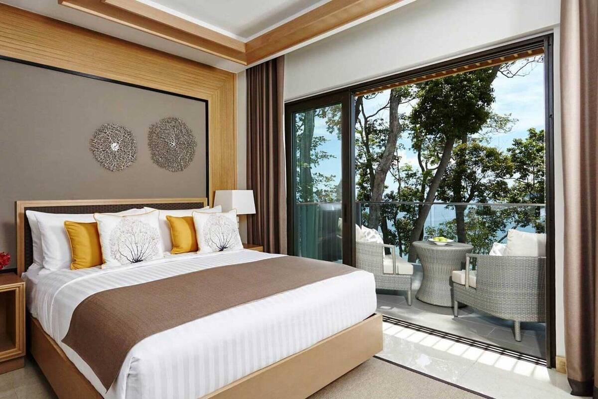 Een moderne hotelkamer met een groot bed, twee nachtkastjes, een balkon met rieten meubilair en een mooi uitzicht.