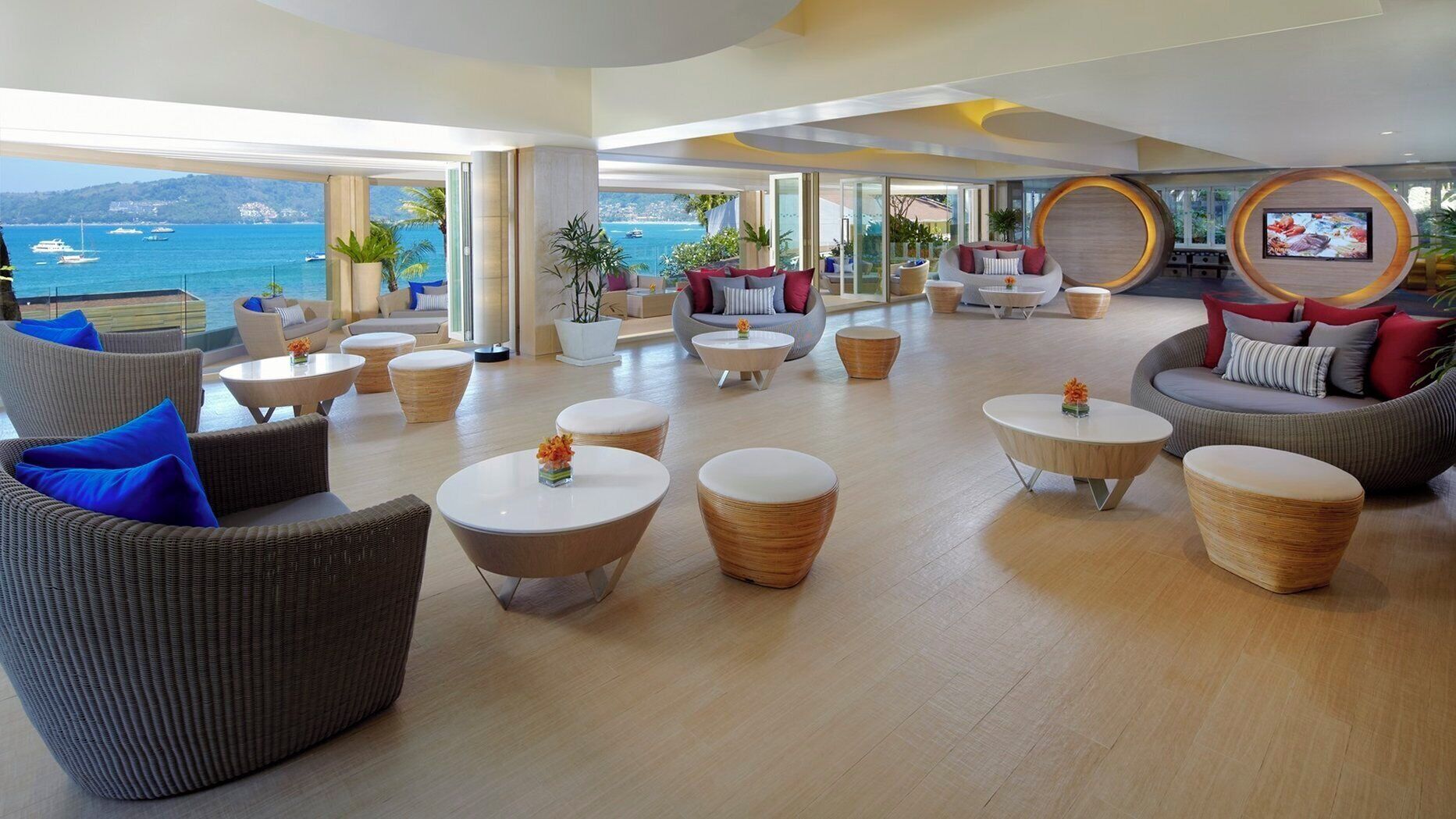 A spacious lounge area with wicker furniture, round tables, and a large TV screen.