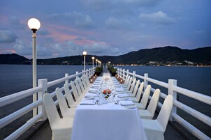 Una larga mesa de comedor lista para una comida, con sillas blancas, mantel blanco y velas a lo largo de la barandilla.