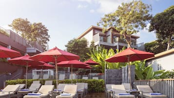 A poolside area with lounge chairs and red umbrellas, surrounded by greenery and a modern building.