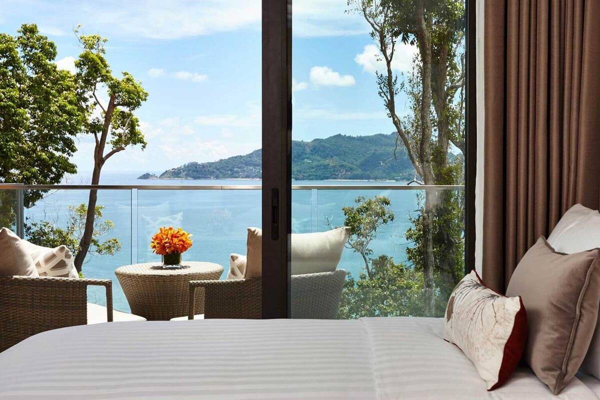 Een hotelkamer met een groot bed, uitzicht op zee en een balkon met rieten meubels en een bloemenvaas.