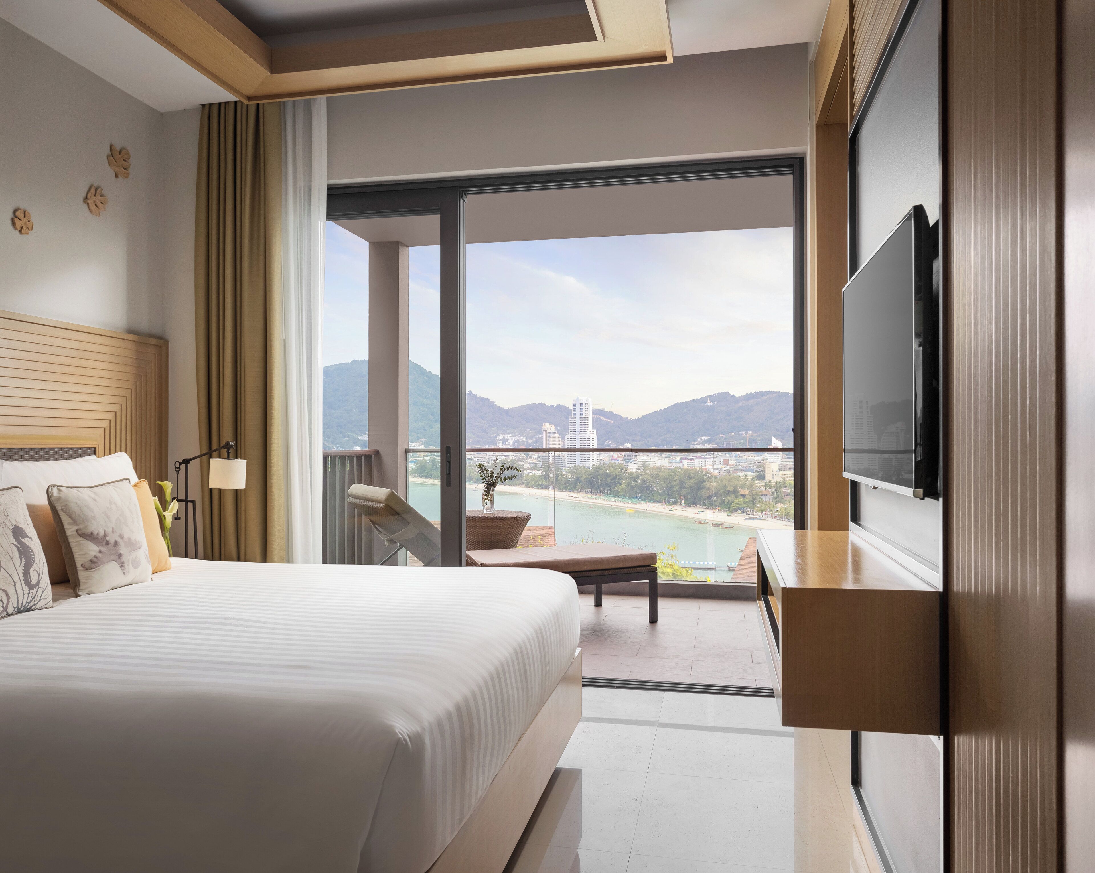 Ein Hotelzimmer mit einem großen Bett, einem Balkon mit Blick auf Strand und Berge sowie einem an der Wand befestigten Flachbildfernseher.