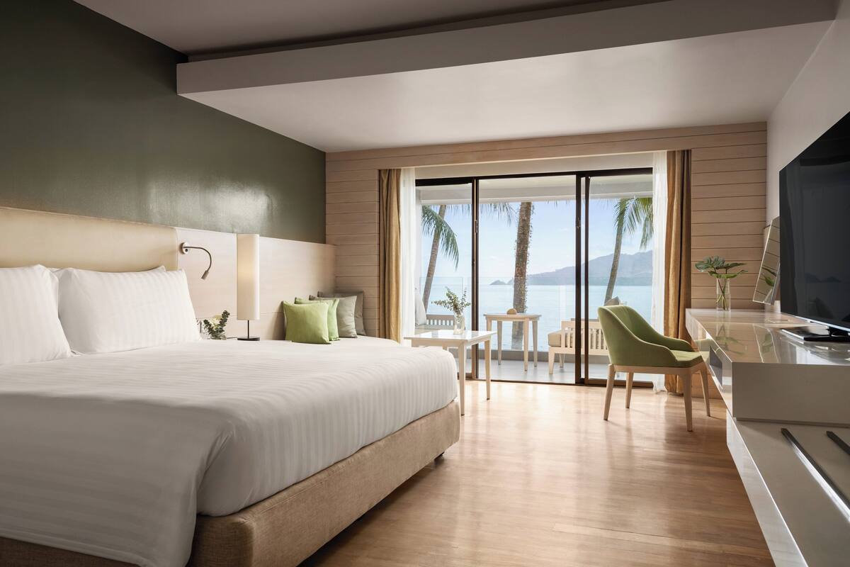 Een moderne hotelkamer met een groot bed, een bureau met televisie en uitzicht op palmbomen en bergen.
