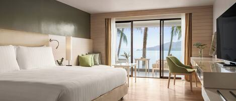 A modern hotel room with a large bed, a desk with a TV, and a view of palm trees and mountains.