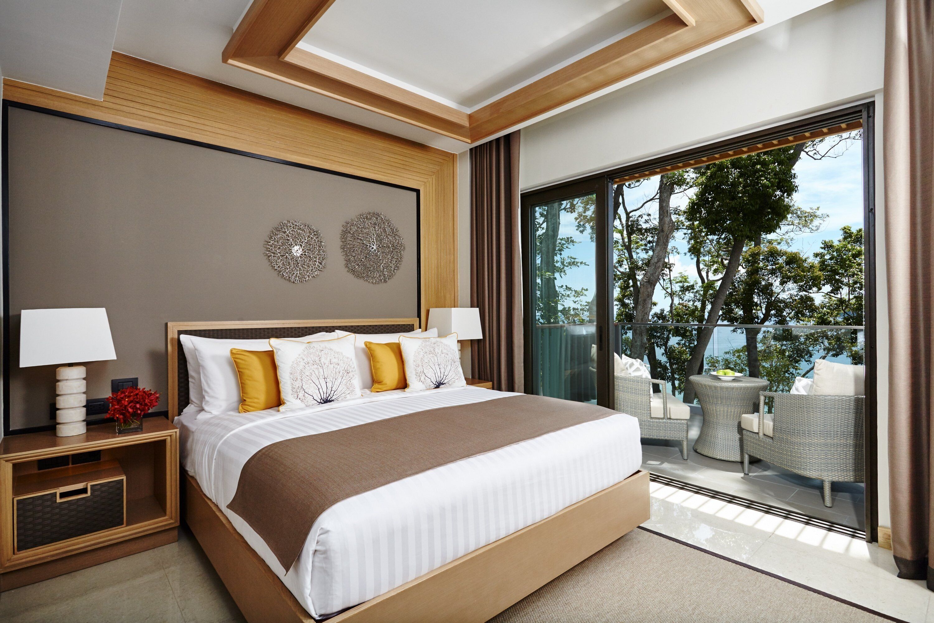 A modern hotel room with a large bed, two wall decor pieces, a nightstand with a lamp, and a balcony with wicker furniture and a view of trees.