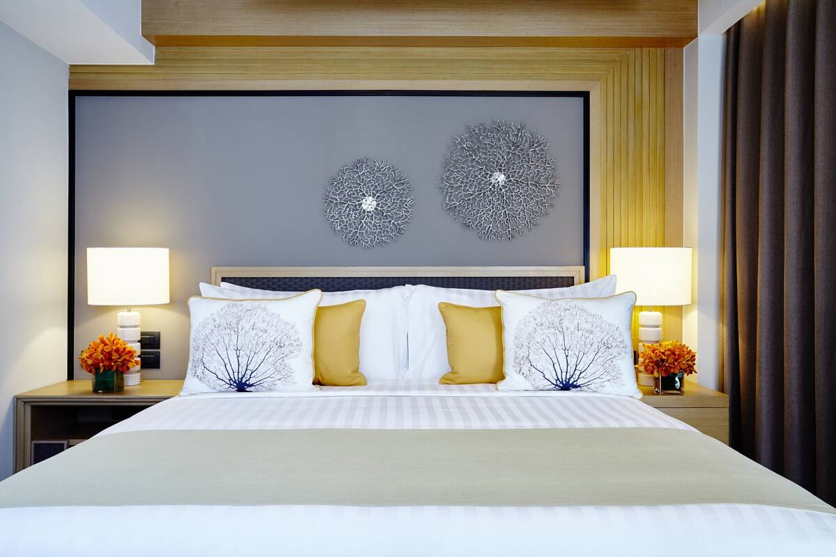 Een net opgemaakt bed met wit en goud linnengoed, twee decoratieve wandpanelen, en twee bedlampen met bloemstukken.