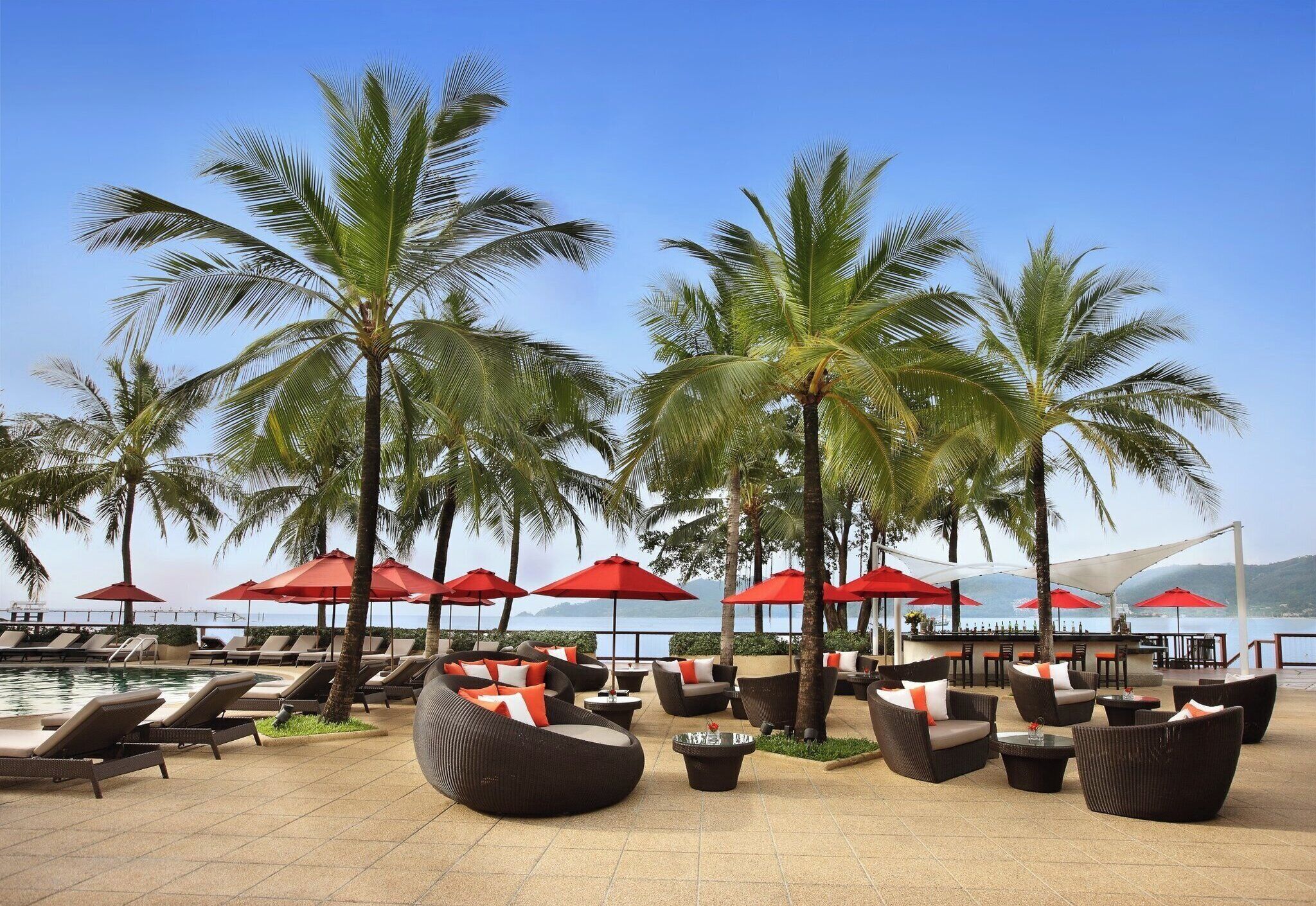 A poolside area with lounge chairs and umbrellas, palm trees, and a view of the sea.