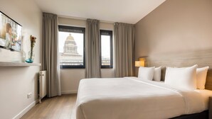 A hotel room with a large bed, a TV on the wall, a window with a view of a building, and a radiator. - Citadines Toison d'Or Brussels (Brussels)