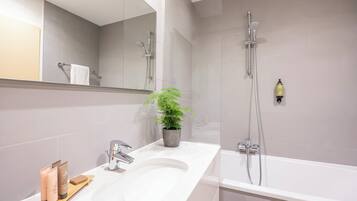 Une salle de bain moderne avec un grand lavabo blanc, un miroir, une douche équipée d’un pommeau de douche à main et une plante en pot.