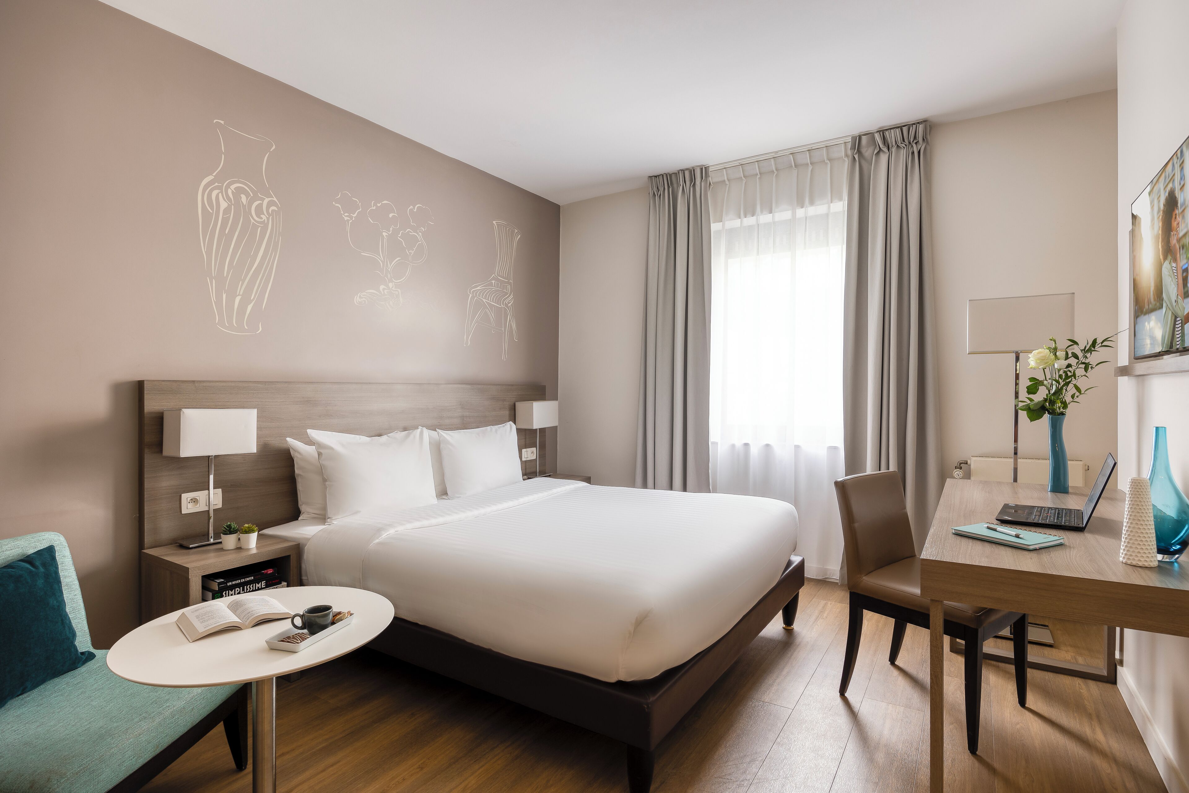 A hotel room with a large bed, a desk with a laptop, a chair, a small table with a book and a cup, and a window with curtains.