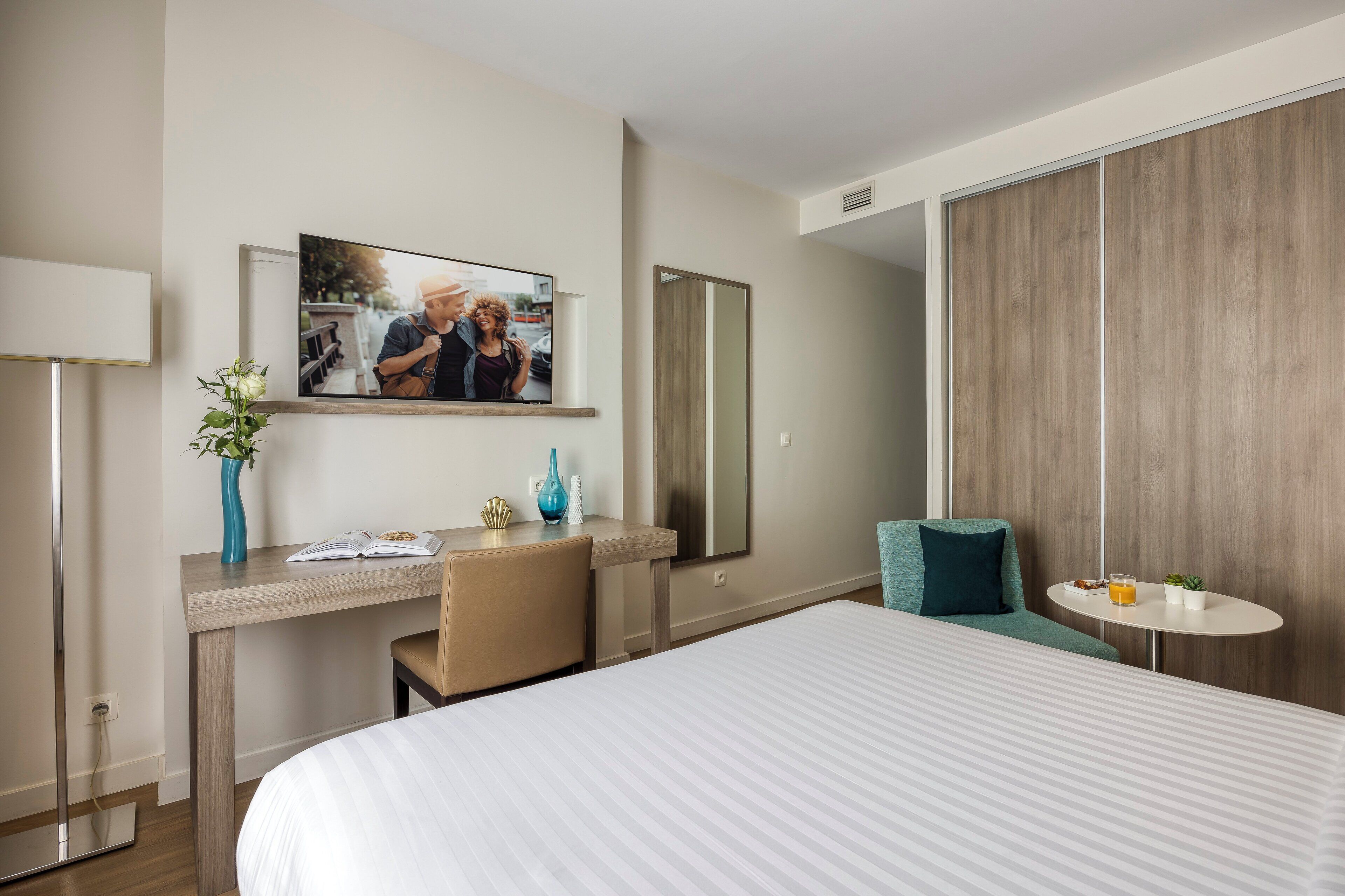 A hotel room with a bed, a desk with a chair, a TV mounted on the wall, and a small table with a plant and a drink.