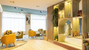 A modern hotel lobby with a wooden floor, yellow armchairs, a central coffee table, and a bookshelf with books and decorative items. - Citadines Toison d'Or Brussels (Brussels)