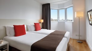 A hotel room with two beds, a large window with a view, a floor lamp, and a nightstand with a book. - Citadines Montparnasse Paris (Paris)