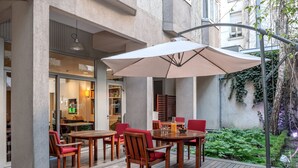An outdoor patio with wooden tables and chairs, a large umbrella, and a view of the building's interior. - Citadines Montparnasse Paris (Paris)