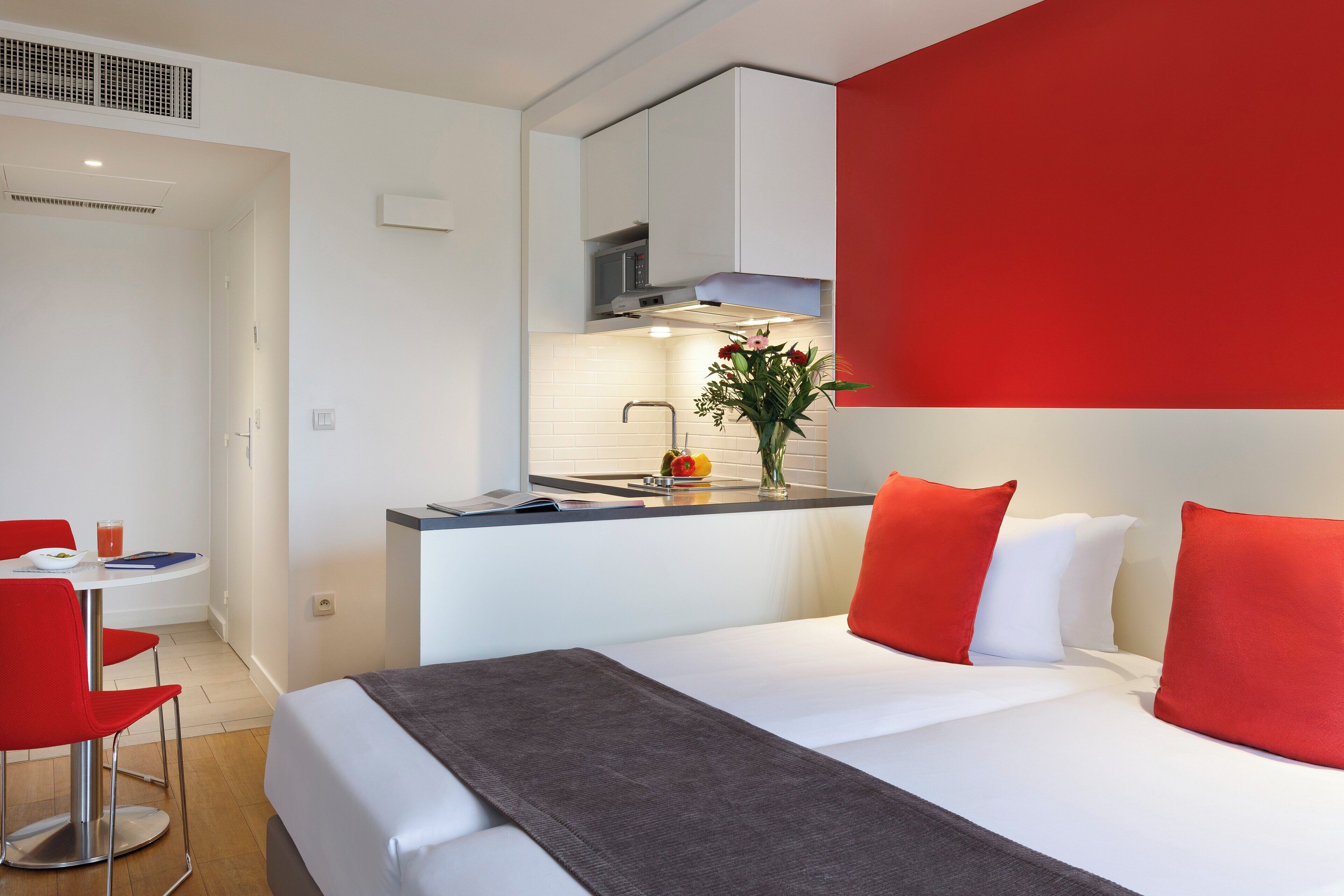 A modern hotel room with a bed, a small kitchenette, a dining area, and a red accent wall.