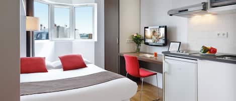 A modern hotel room with a bed, a desk, a chair, a small kitchen area, and a window with a city view.