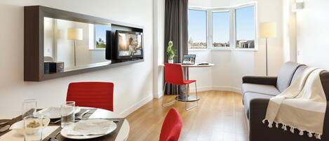 Habitación de hotel moderna con comedor, sala de estar con sofá, televisor montado en la pared y ventanas amplias con vistas a la ciudad.