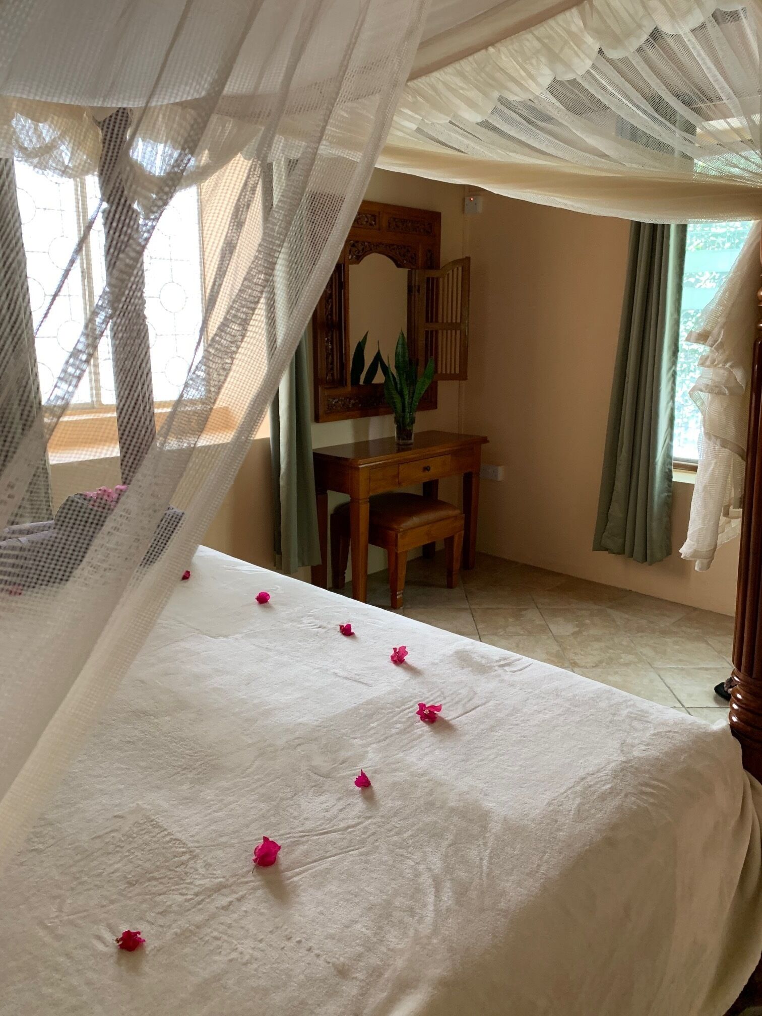 A room with a bed covered in white linens, a wooden desk with a mirror, and a potted plant.