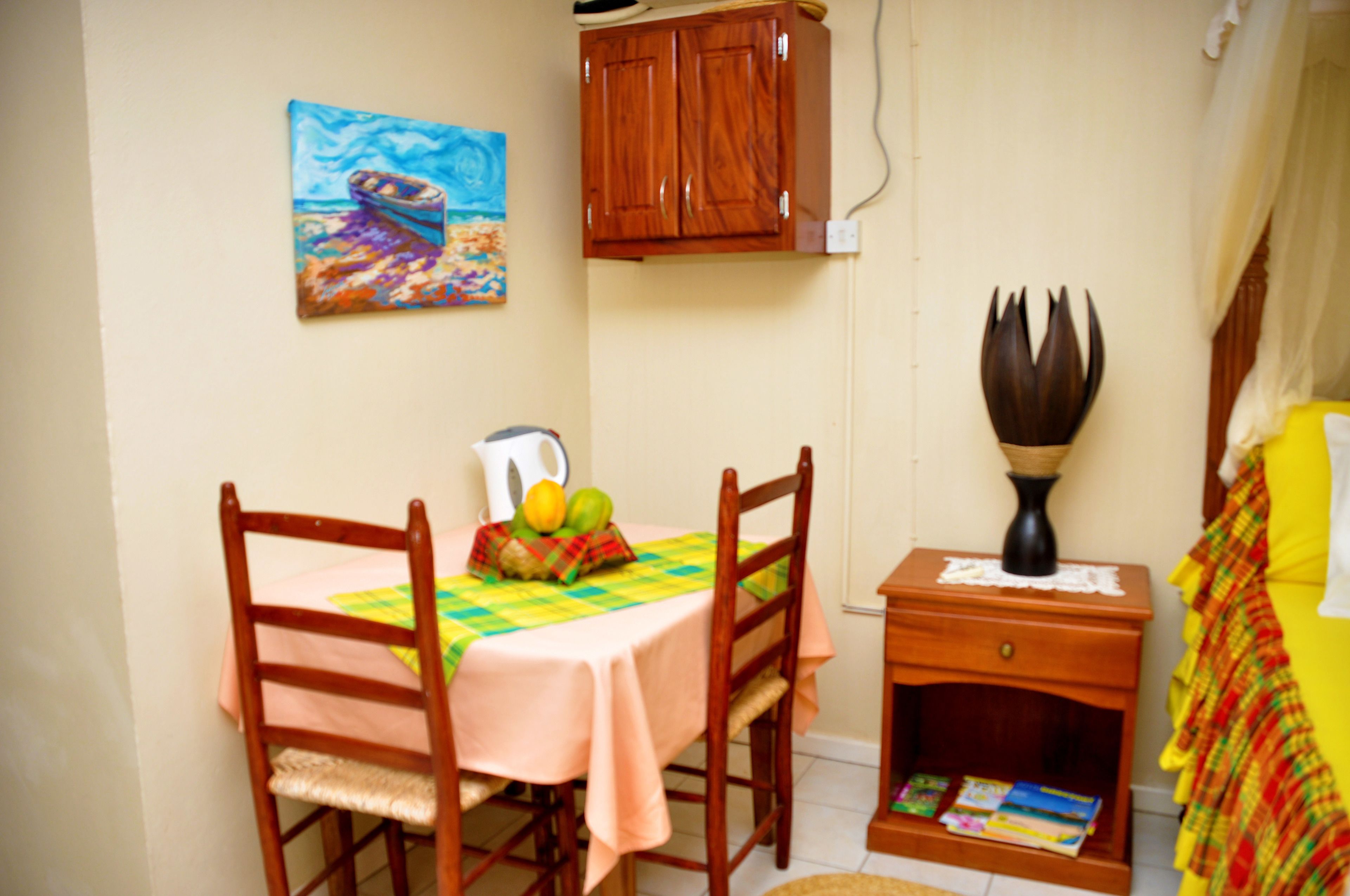 A small dining area with a table set for two, a wooden cabinet above, and a bedside table with a lamp.