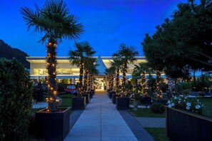 A pathway lined with palm trees and planters, leading to a building with a modern design and a pointed roof. - Hotel Egerkingen (Egerkingen)