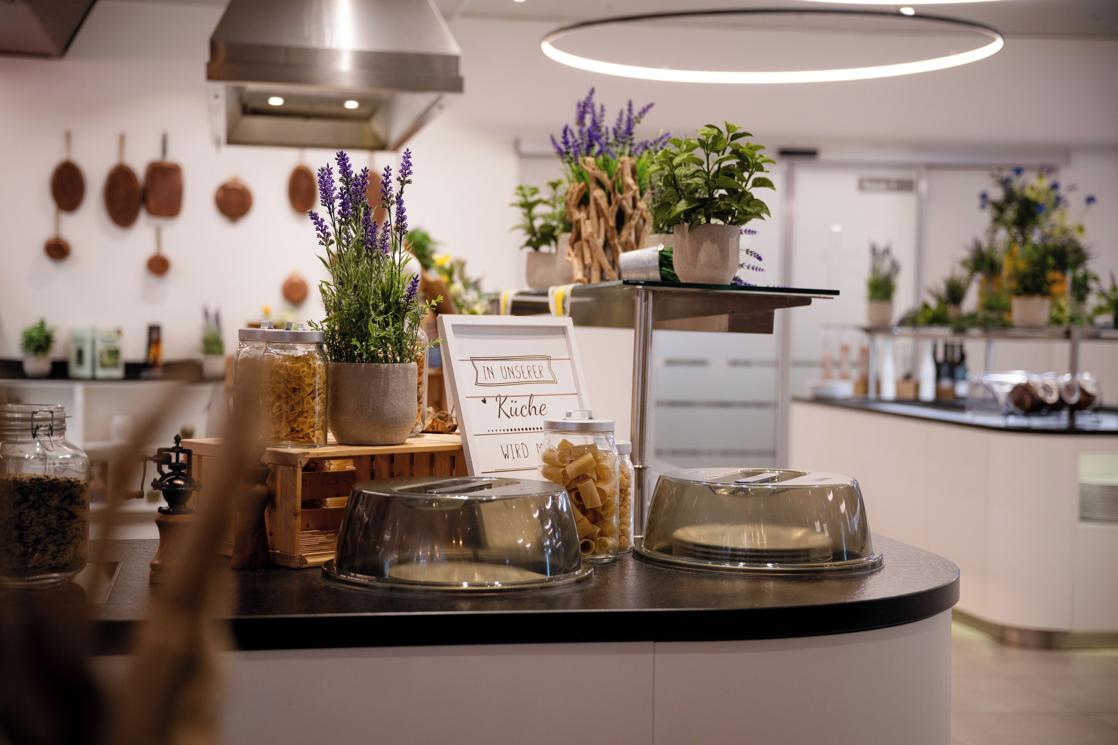 A kitchen counter with jars of pasta, a sign reading "IN INSEPER Kueche," and various potted plants.