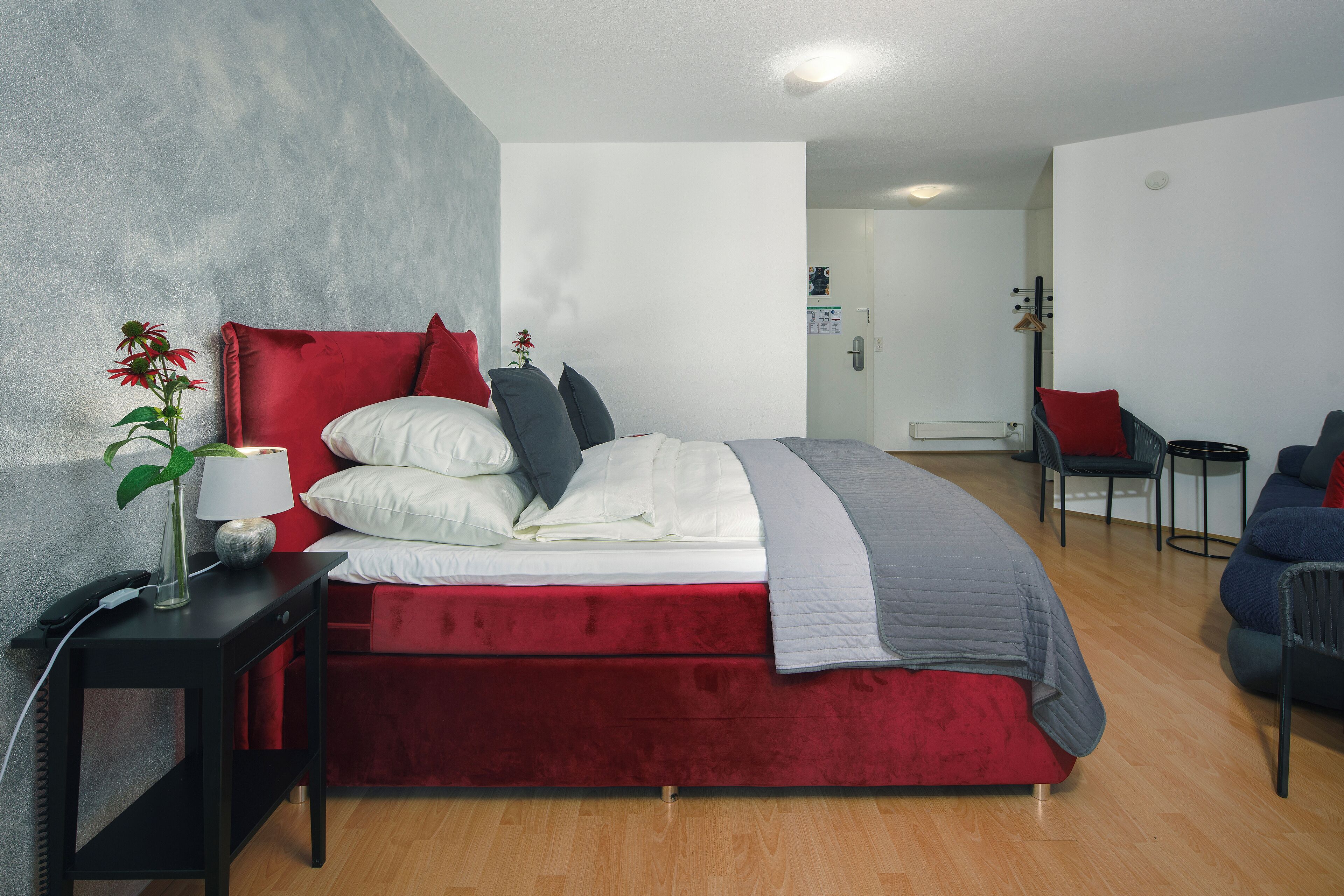 Un dormitorio moderno con cama roja, ropa de cama blanca y gris, una mesita de noche pequeña con una lámpara y una planta.