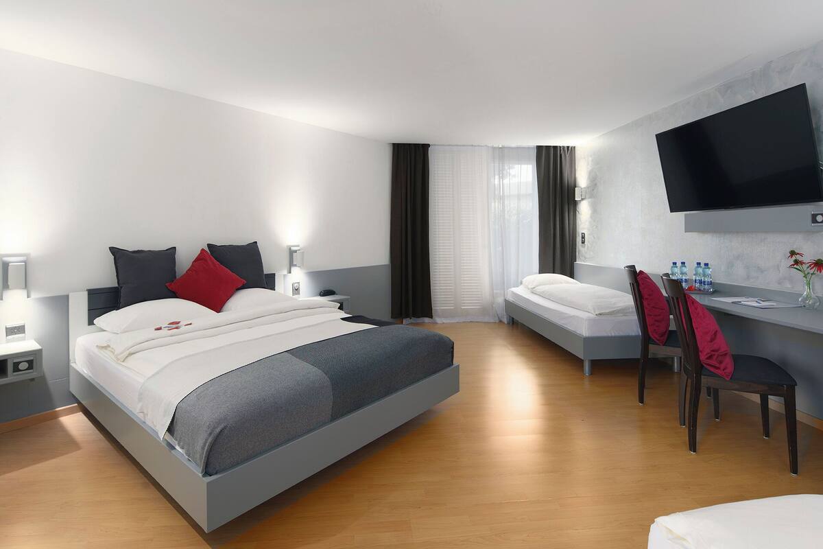 A modern hotel room with a bed, a desk, a chair, a TV, and a window with curtains.