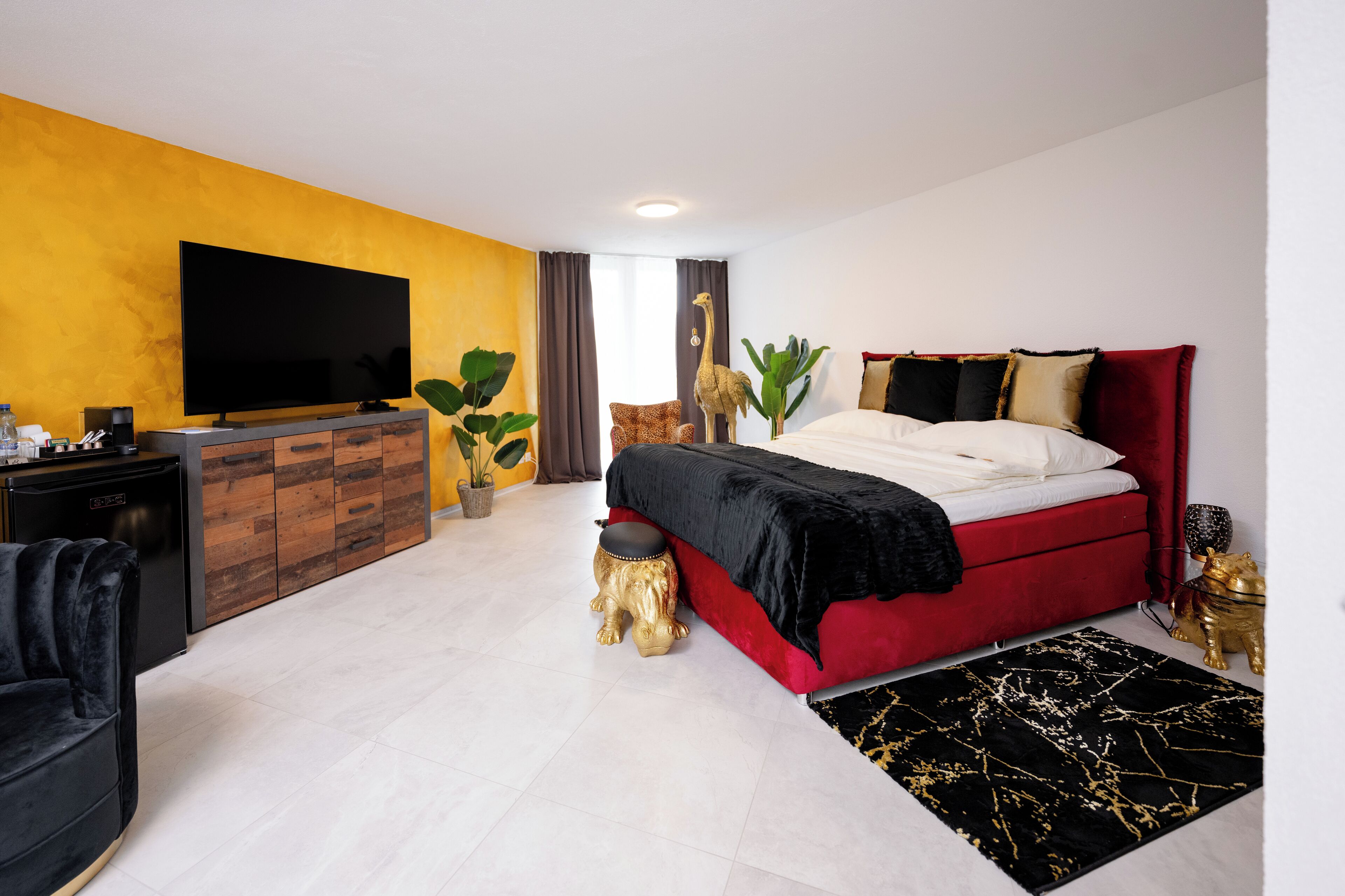 Moderni hotellihuone, jossa on suuri litteänäyttöinen televisio, punainen sänky, mustavalkoinen ja kultainen kuvioitu matto sekä puinen kaappi, jossa on kasvi.