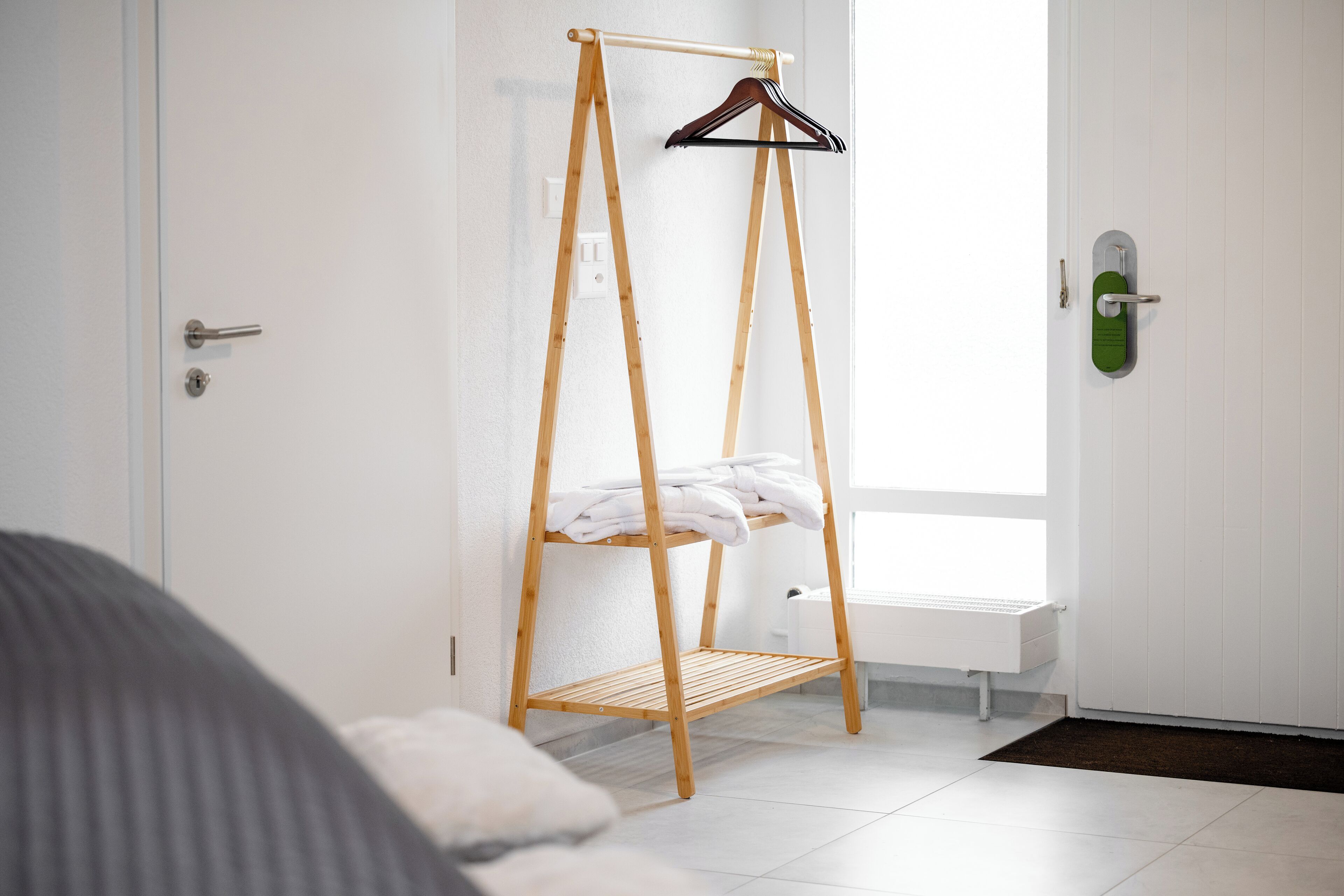 Habitación con un perchero de madera junto a una puerta blanca, una cama con edredón blanco y una ventana que deja entrar luz natural.
