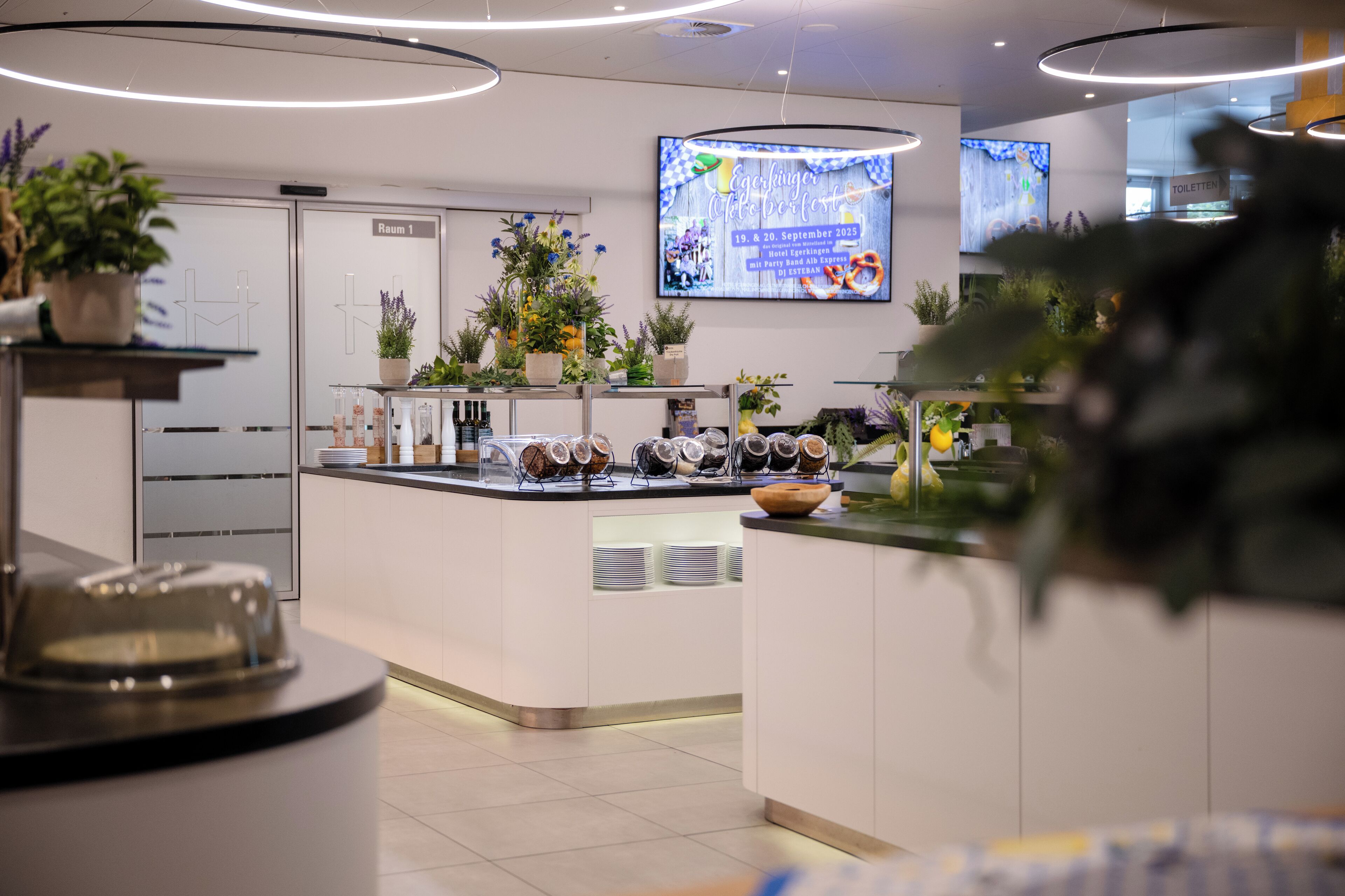 Une cuisine d’hôtel moderne, dotée d’un îlot central, de plantes en pot et d’un écran numérique affichant des informations sur les événements.