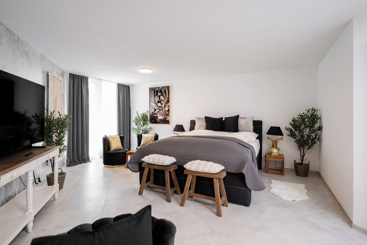 A modern bedroom with a large bed, two stools, a TV, and a dining table.