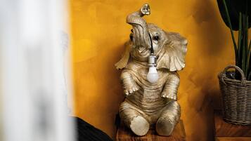 Une sculpture décorative représentant un éléphant, avec une ampoule sur la tête, posée sur un tabouret en bois devant un mur jaune.