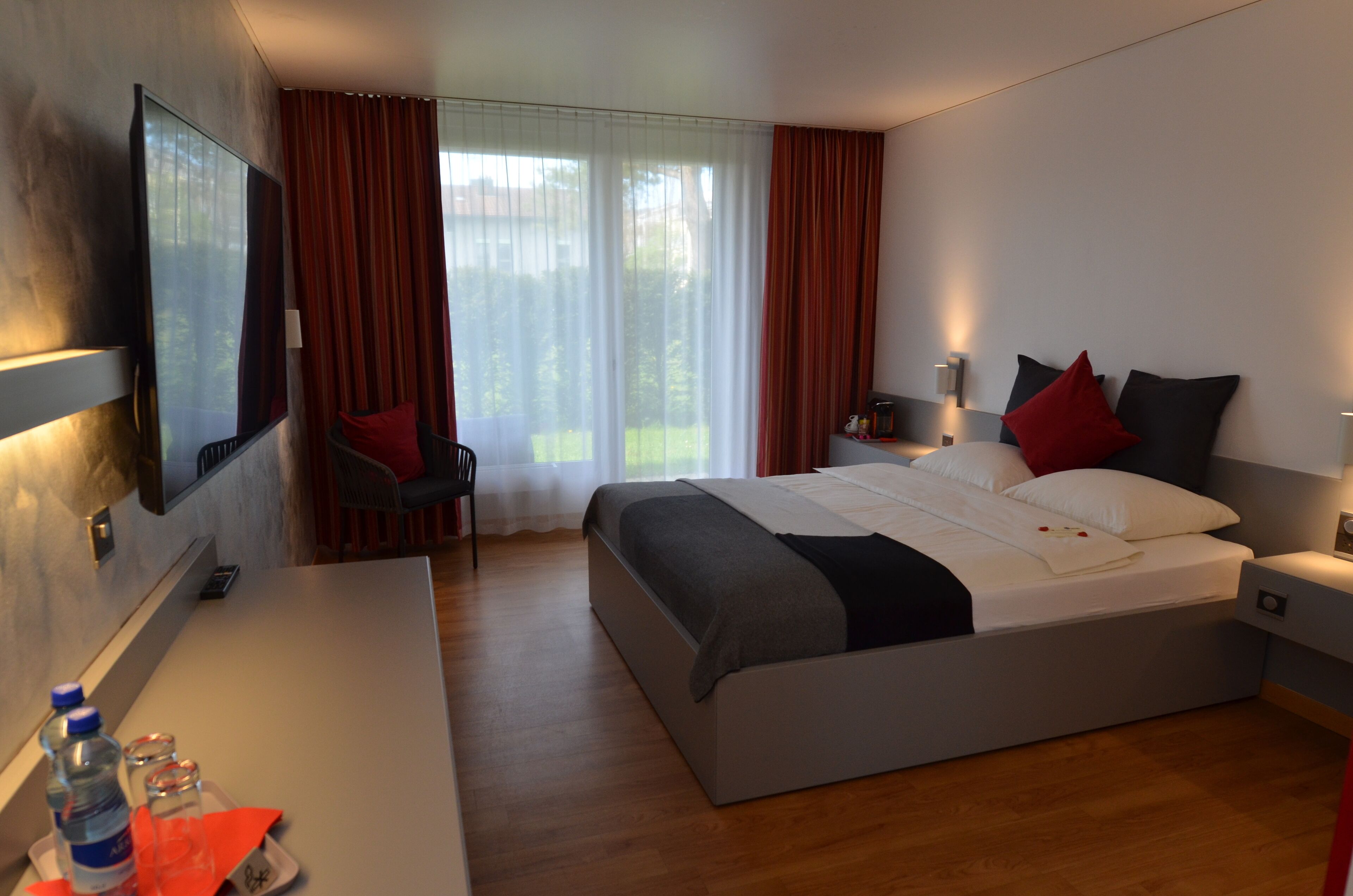Ein modernes Hotelzimmer mit einem großen Bett, einem Schreibtisch mit Stuhl, einem an der Wand montierten Fernseher und einem Fenster mit Vorhängen.
