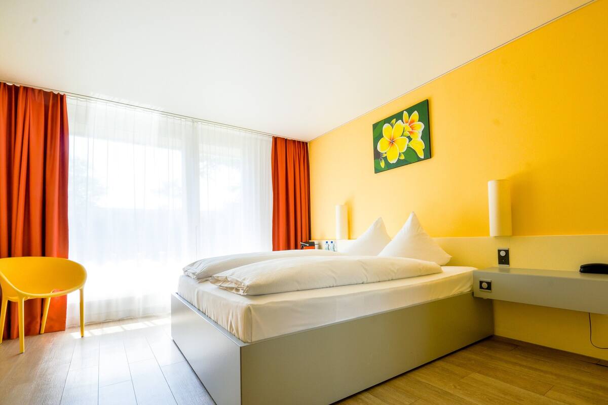 A modern hotel room with a large bed, a bright yellow wall, and a window with orange curtains.