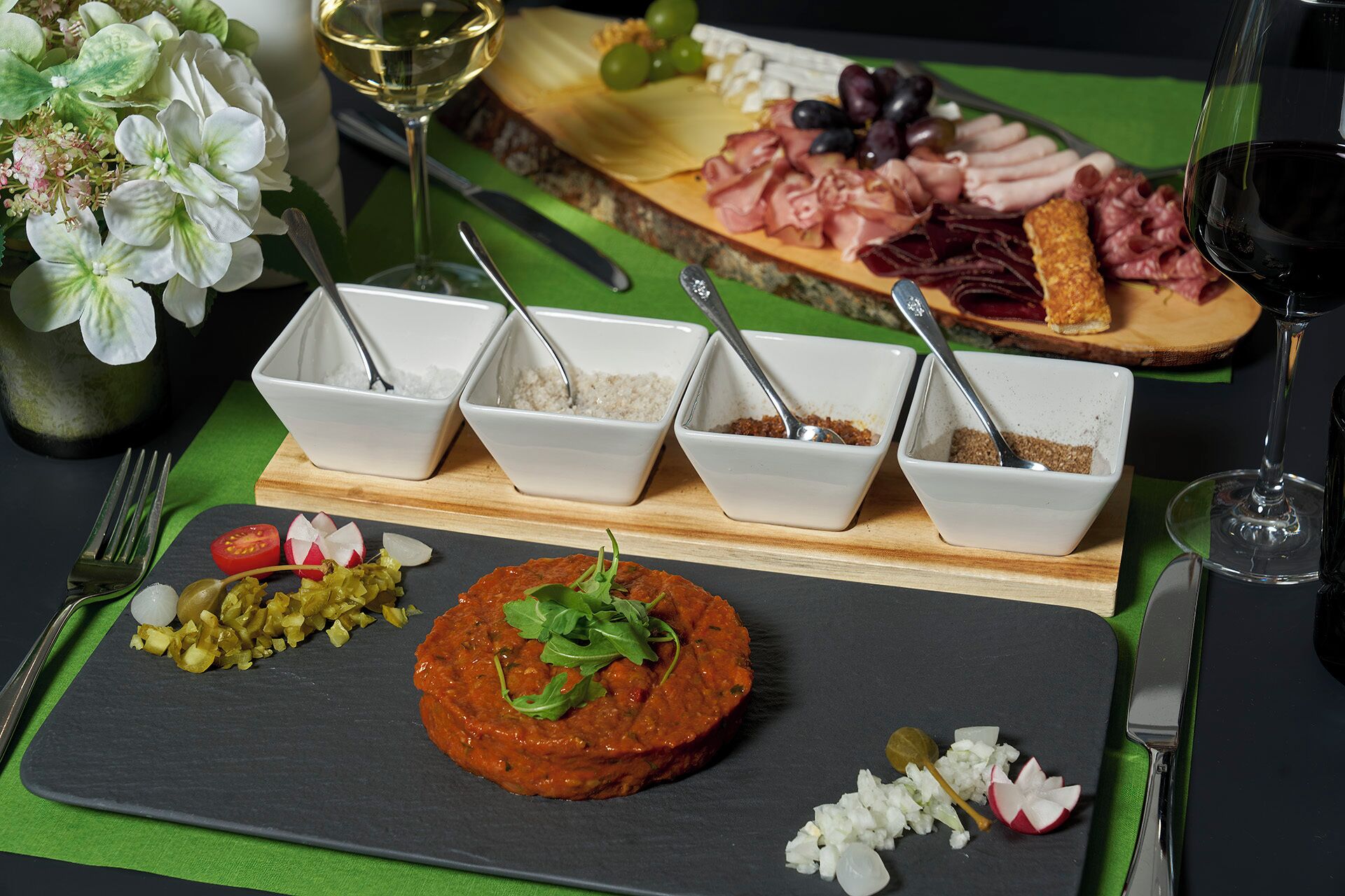Una tabla de fiambres con diversos embutidos, quesos y acompañamientos sobre una tabla de madera, acompañada de recipientes con salsas y una copa de vino.