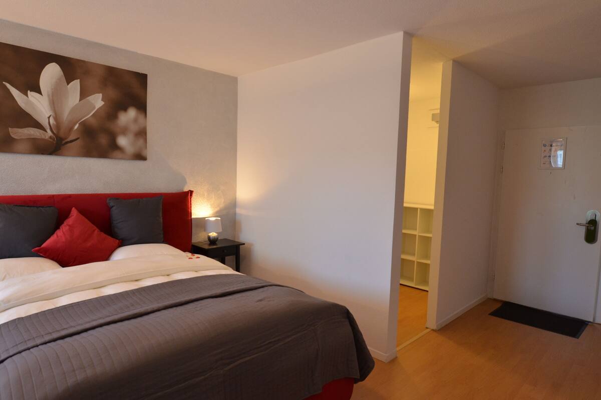 A hotel room with a bed, a nightstand, a lamp, and a door leading to another room.