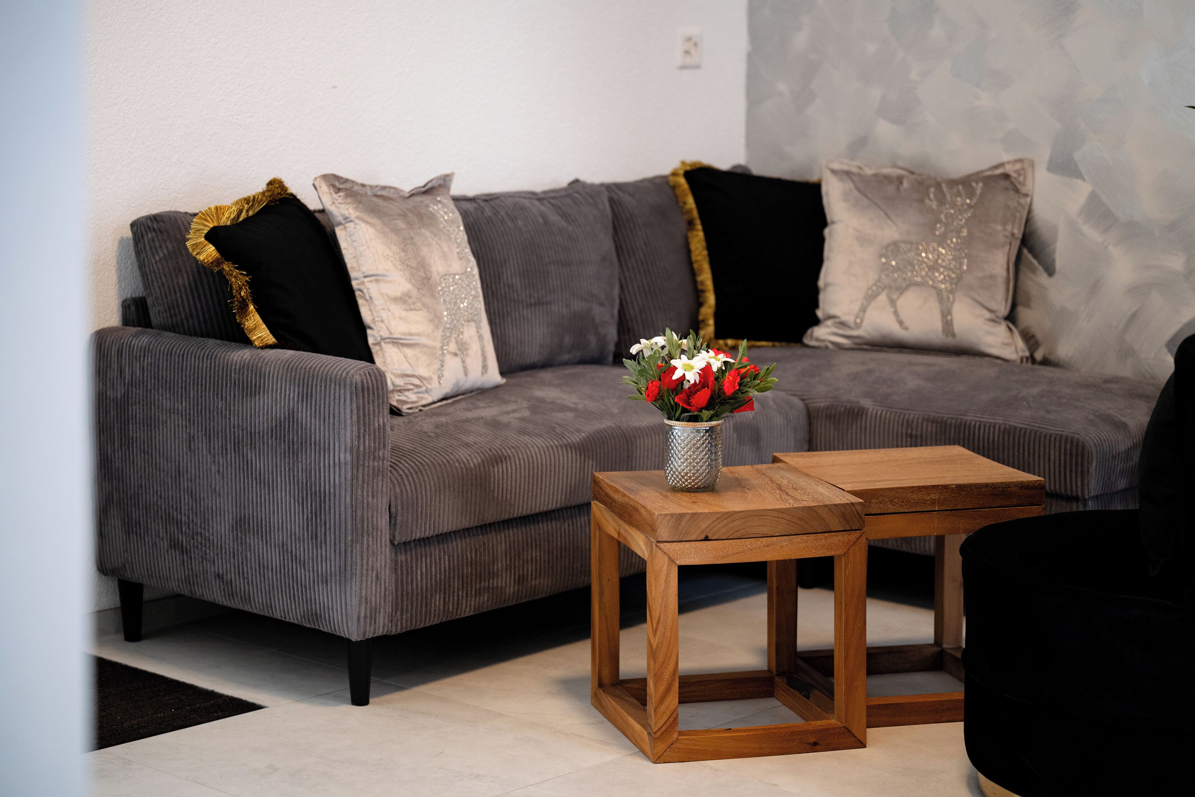 A grey sofa with black and gold pillows, a wooden side table, and a vase with red flowers.