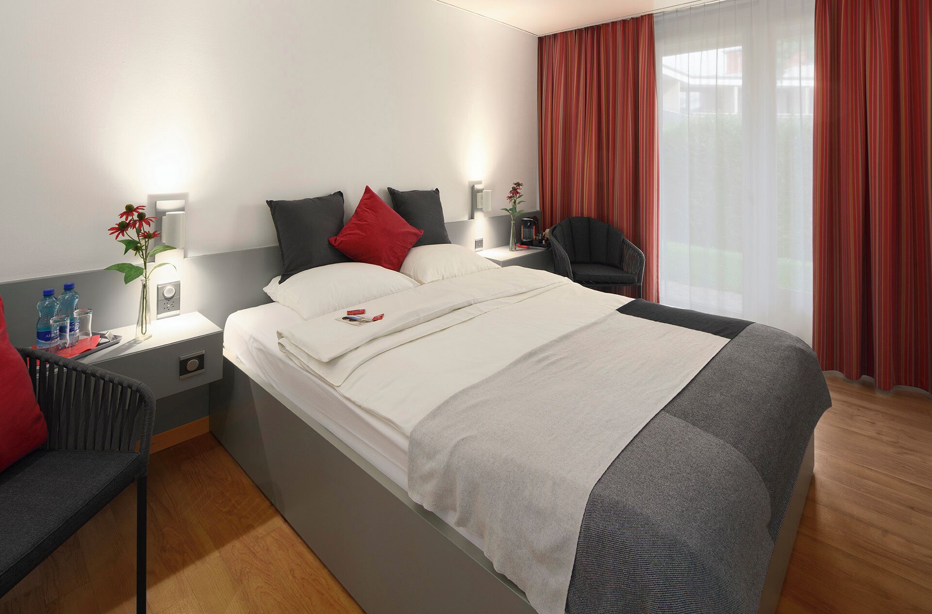A modern hotel room with a neatly made bed, a bedside table with a lamp, a chair, and a view of the outdoors through sheer curtains.