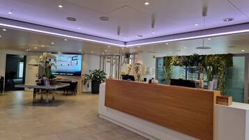 A modern reception area with a wooden reception desk, a large screen displaying information, and a green wall with plants.