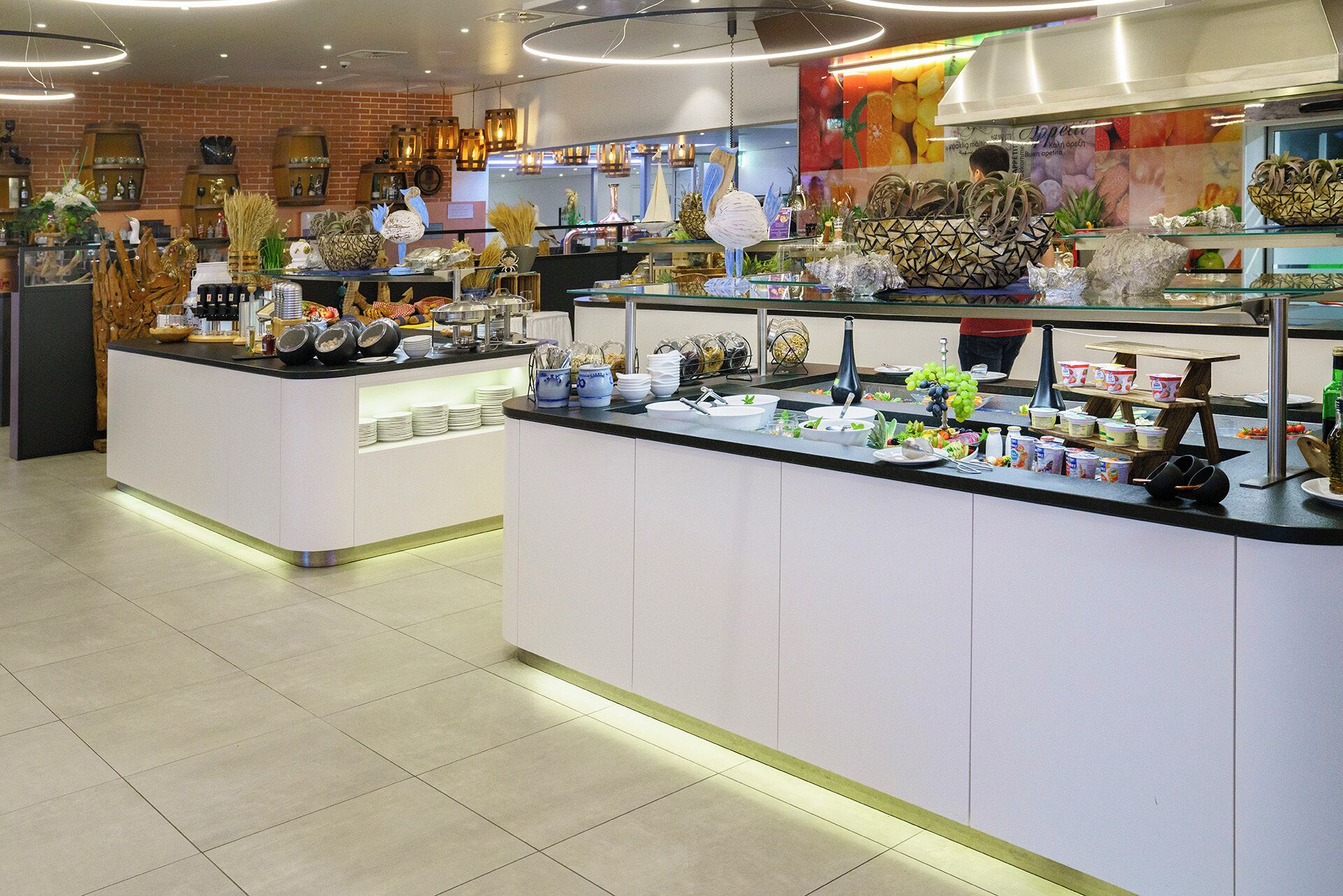 Cafetería moderna y bien iluminada con pared de ladrillo, mostrando una variedad de alimentos y bebidas en mostradores de vidrio.