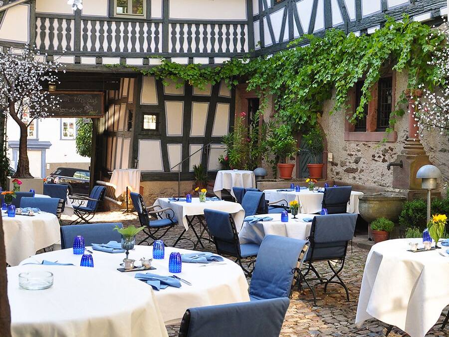 An outdoor dining area with tables and chairs, surrounded by greenery and a building with exposed timber framing.