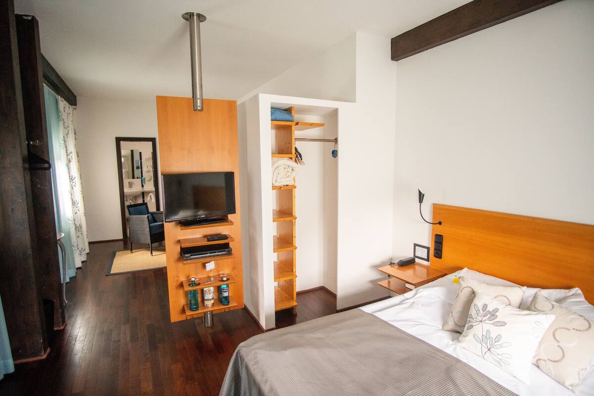 Quarto de hotel com uma cama, televisão, guarda-roupa de madeira e uma pequena escrivaninha com abajur.
