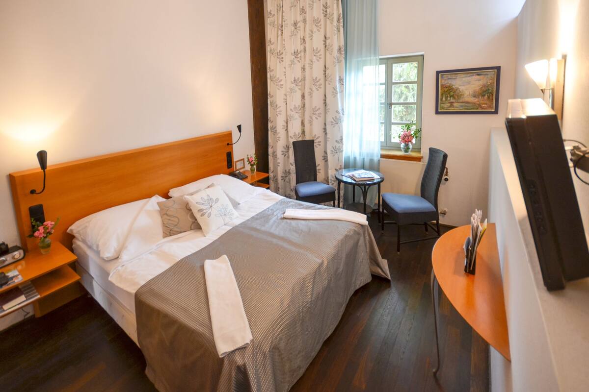 Quarto de hotel com cama, escrivaninha, cadeira, mesinha, janela com cortinas e um quadro na parede.