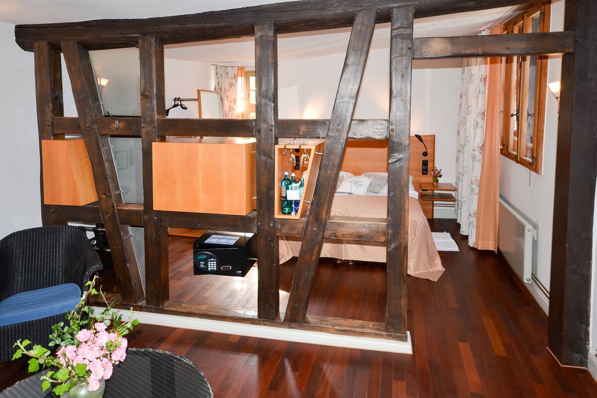 Quarto com vigas de madeira aparentes, cama com cabeceira de madeira, uma mesinha com abajur e uma cadeira.