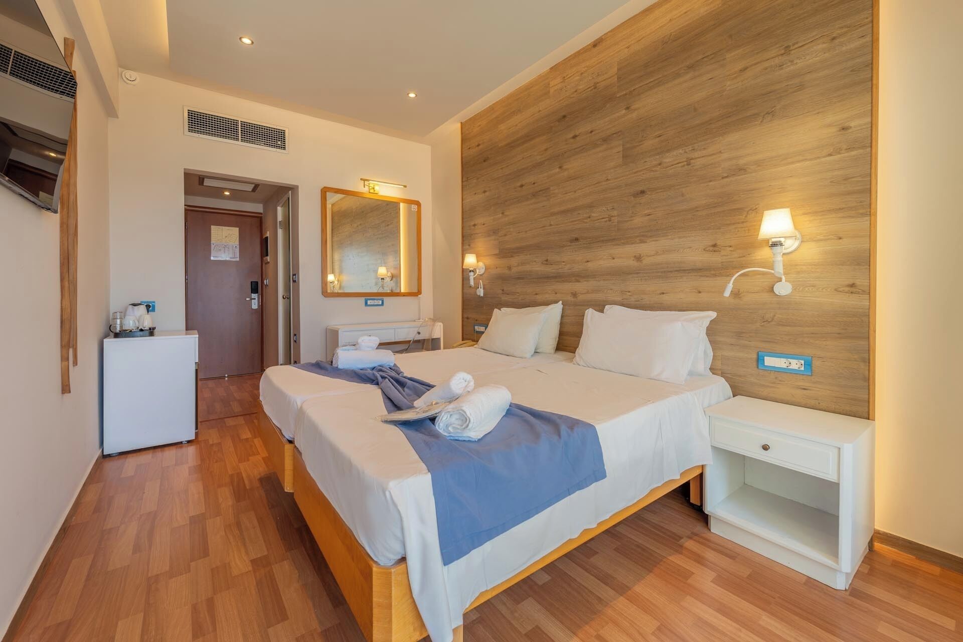 Una habitación de hotel con cabecera de madera, cama con sábanas blancas, mesita de noche, espejo y un escritorio pequeño con lámpara.
