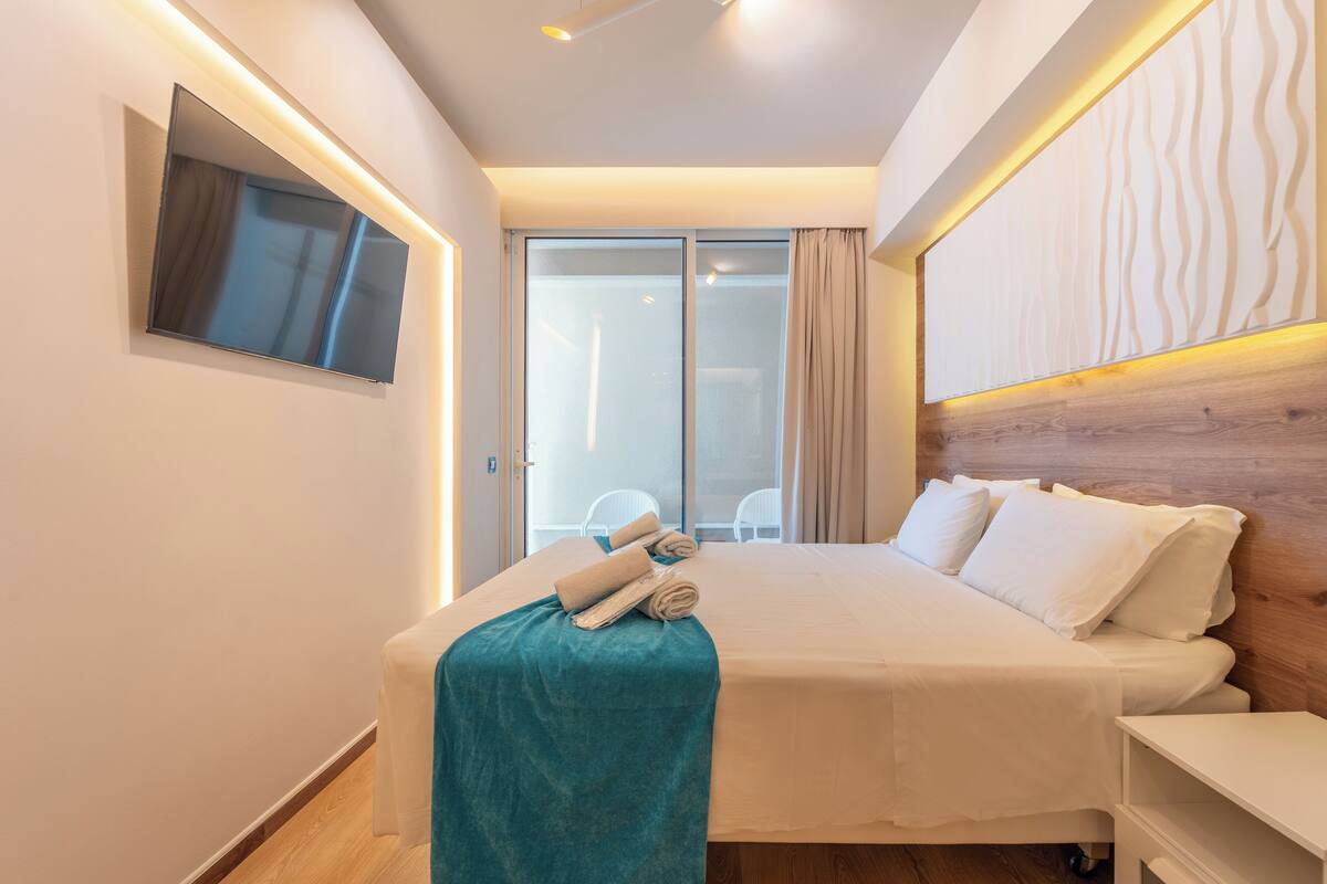 A modern hotel room with a large bed, a flat-screen TV mounted on the wall, and a sliding glass door leading to a balcony.
