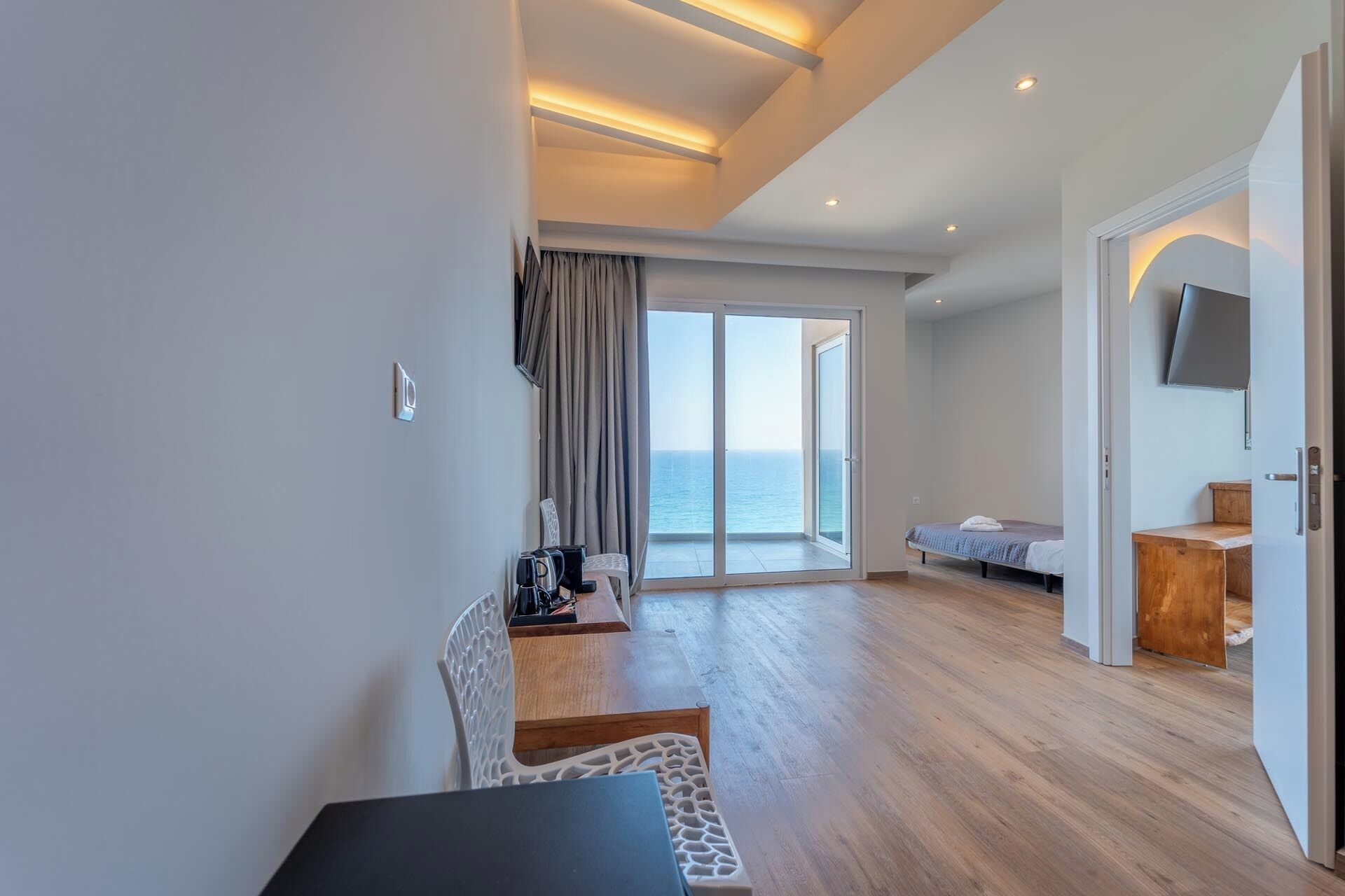 A modern hotel room with a wooden floor, a bed, a desk, and a chair. There is a sliding glass door leading to a balcony with a view of the sea.