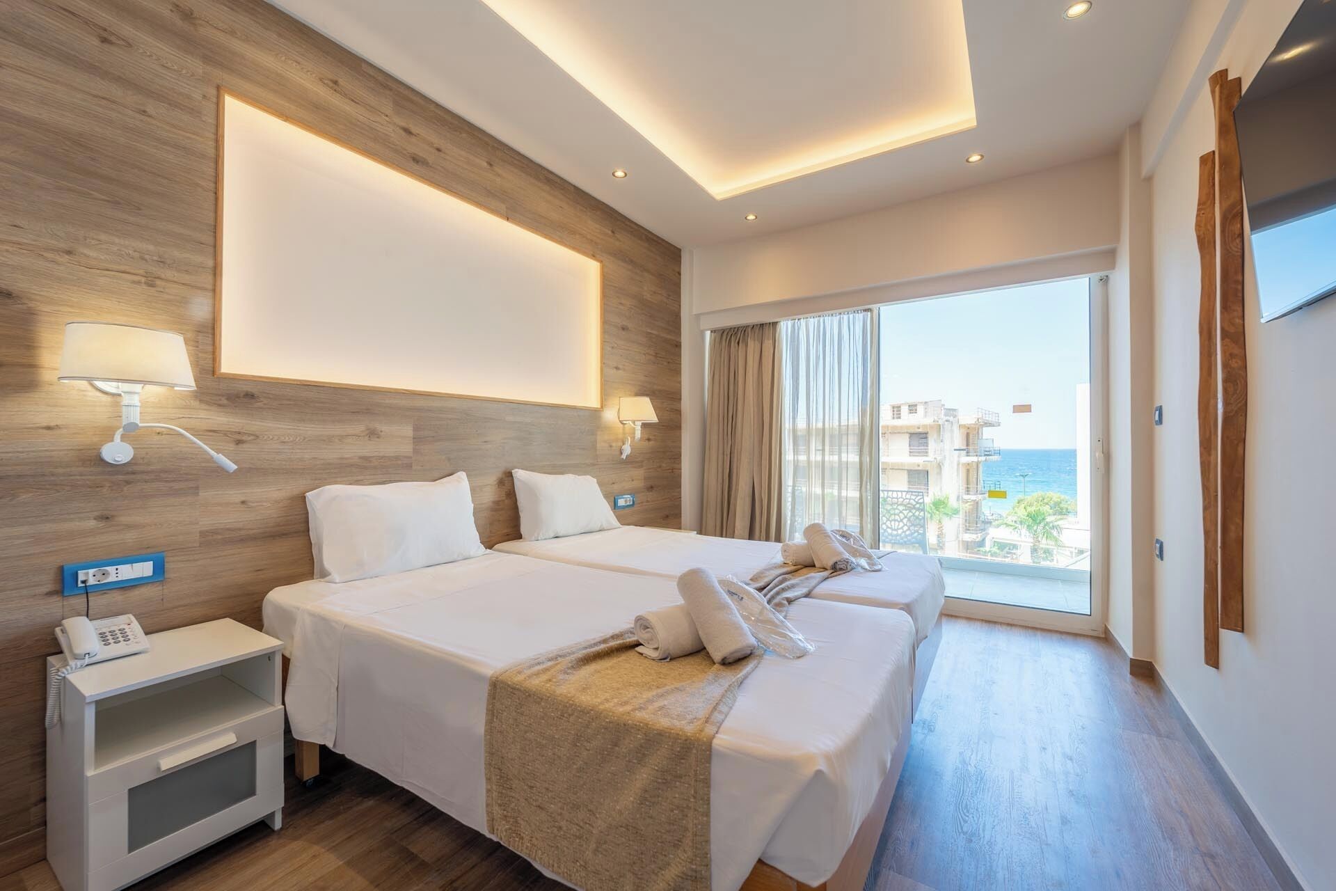 A hotel room with two beds, a bedside table with a phone, a wall-mounted lamp, and a view of the sea.