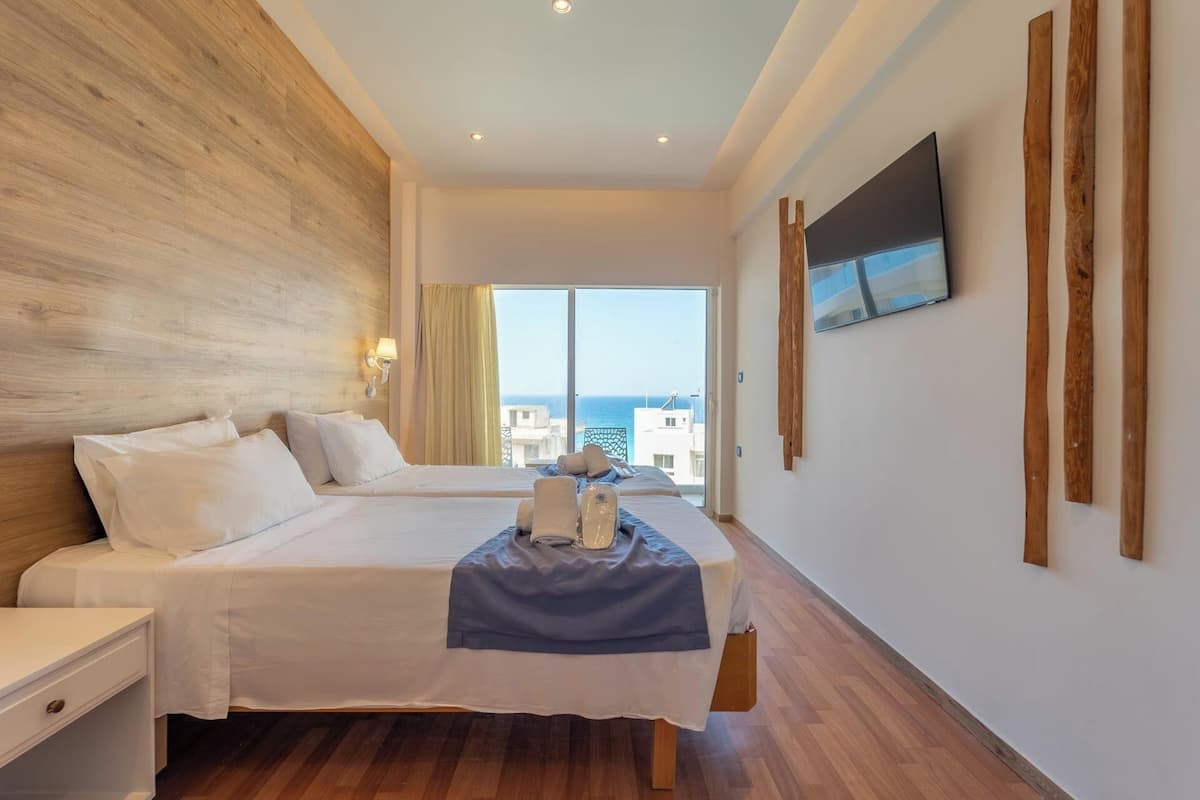 A hotel room with a large bed, a smaller bed, a TV mounted on the wall, and a view of the sea.